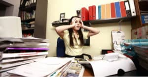 Une étudiante se prend la tête à deux mains devant une pile de documents en désordre qui encombrent son bureau